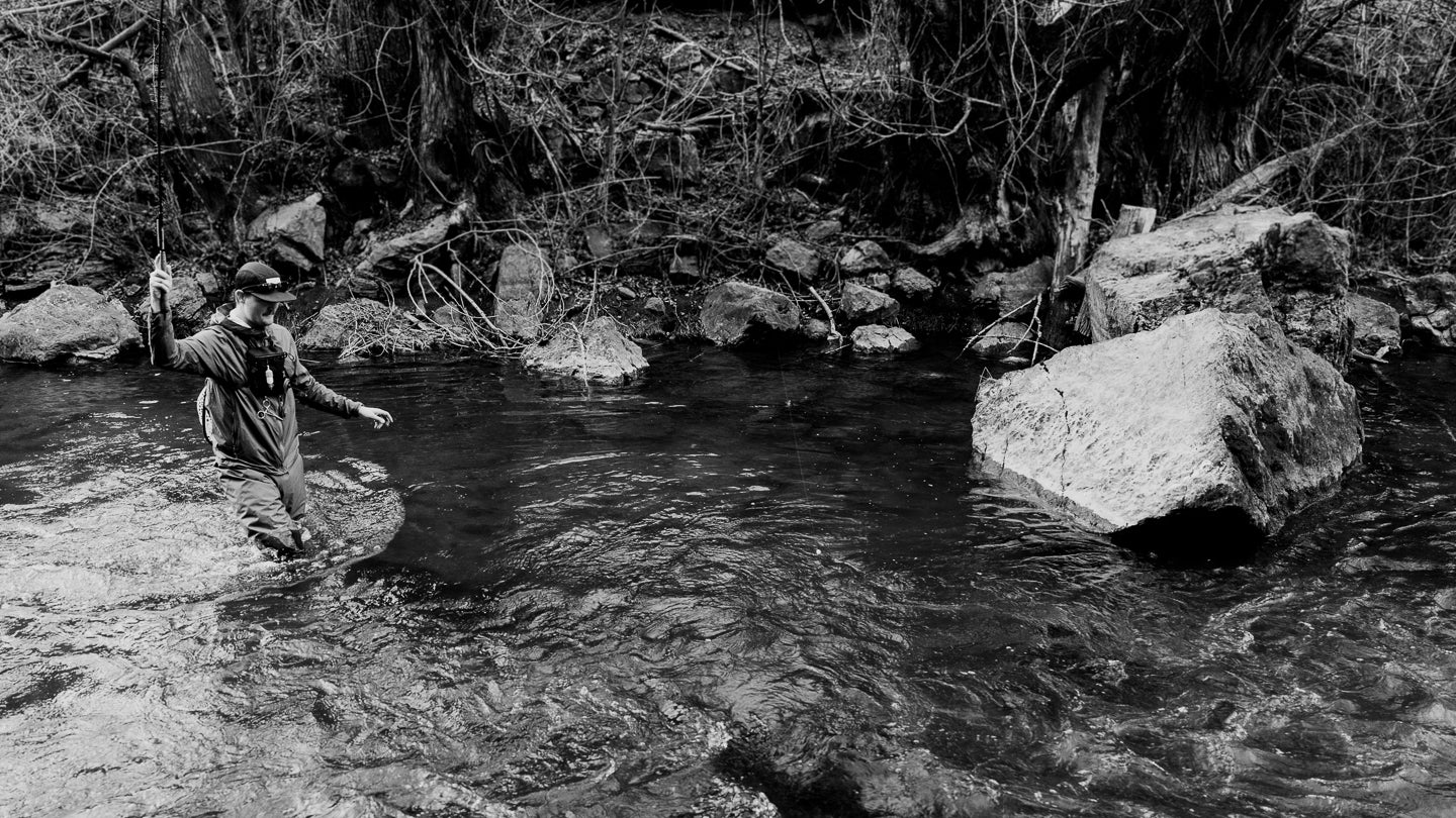 Person fishing in a stream with rocks and trees in the background